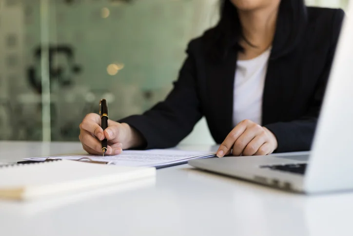 Person signing document at desk with laptop and notebook.